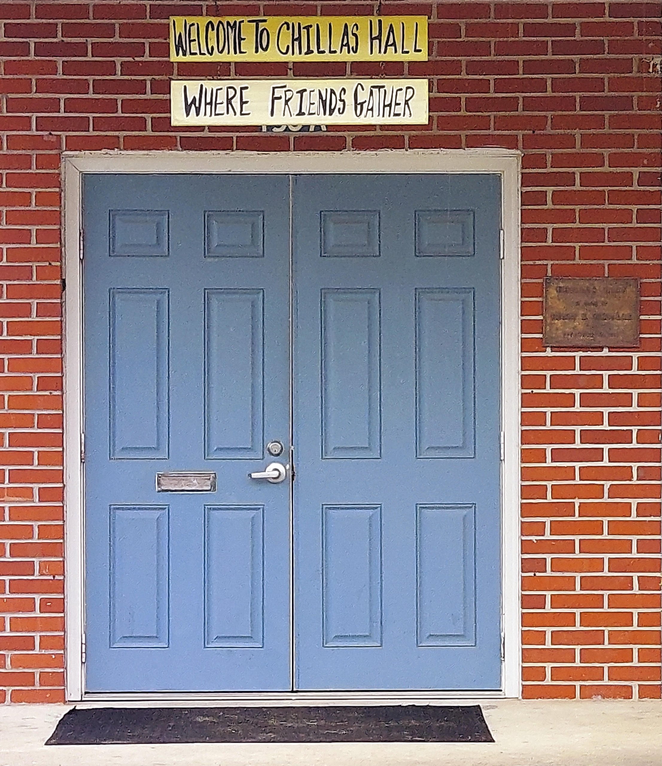 Blue door on red brick building with sign above reading "Welcome to Chillas Hall where friends gather"