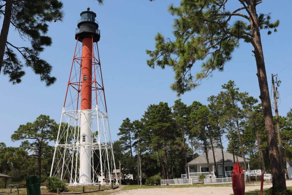 Red and white iron lighthouse tower with black top sits in a park surrounded by green pine trees