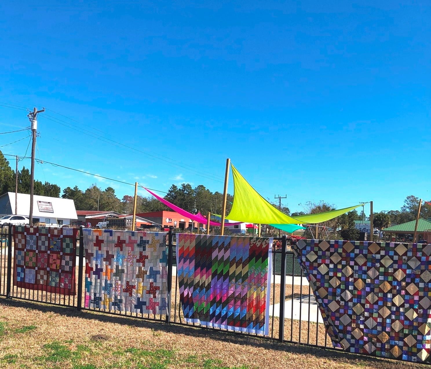Colorful quilts hang on a chain link fence at a city park.
