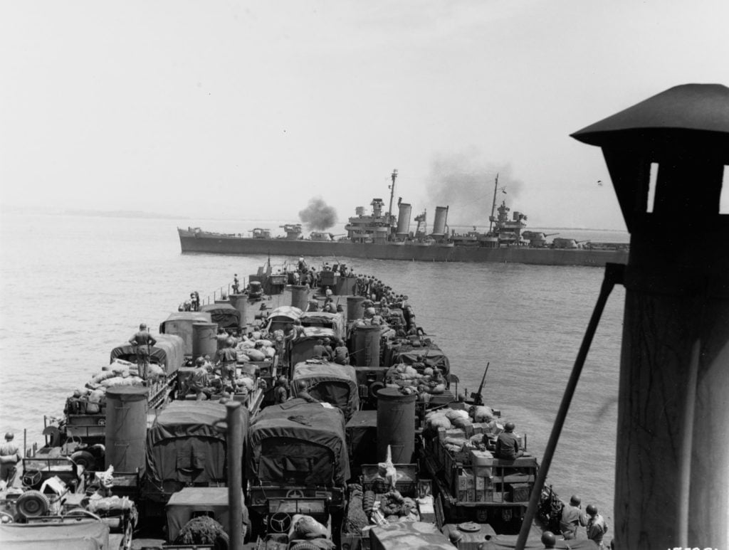 A US WWII ship full of army trucks on the deck fires on another ship