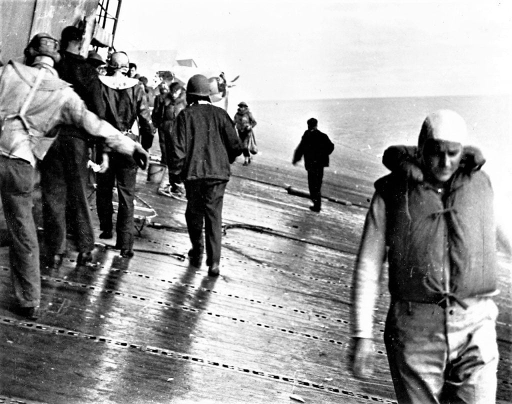 Sailors walk along a WWII ship flight deck while it is listing to the side