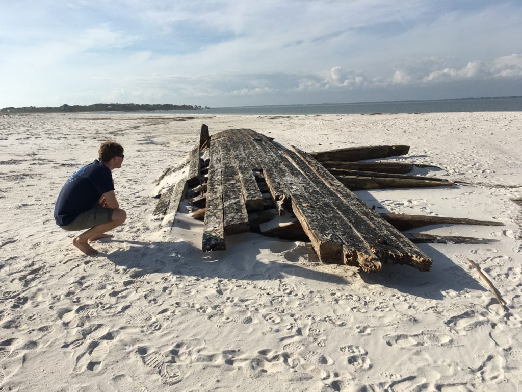 Man kneels beside a portion of a shipwreck on the sand
